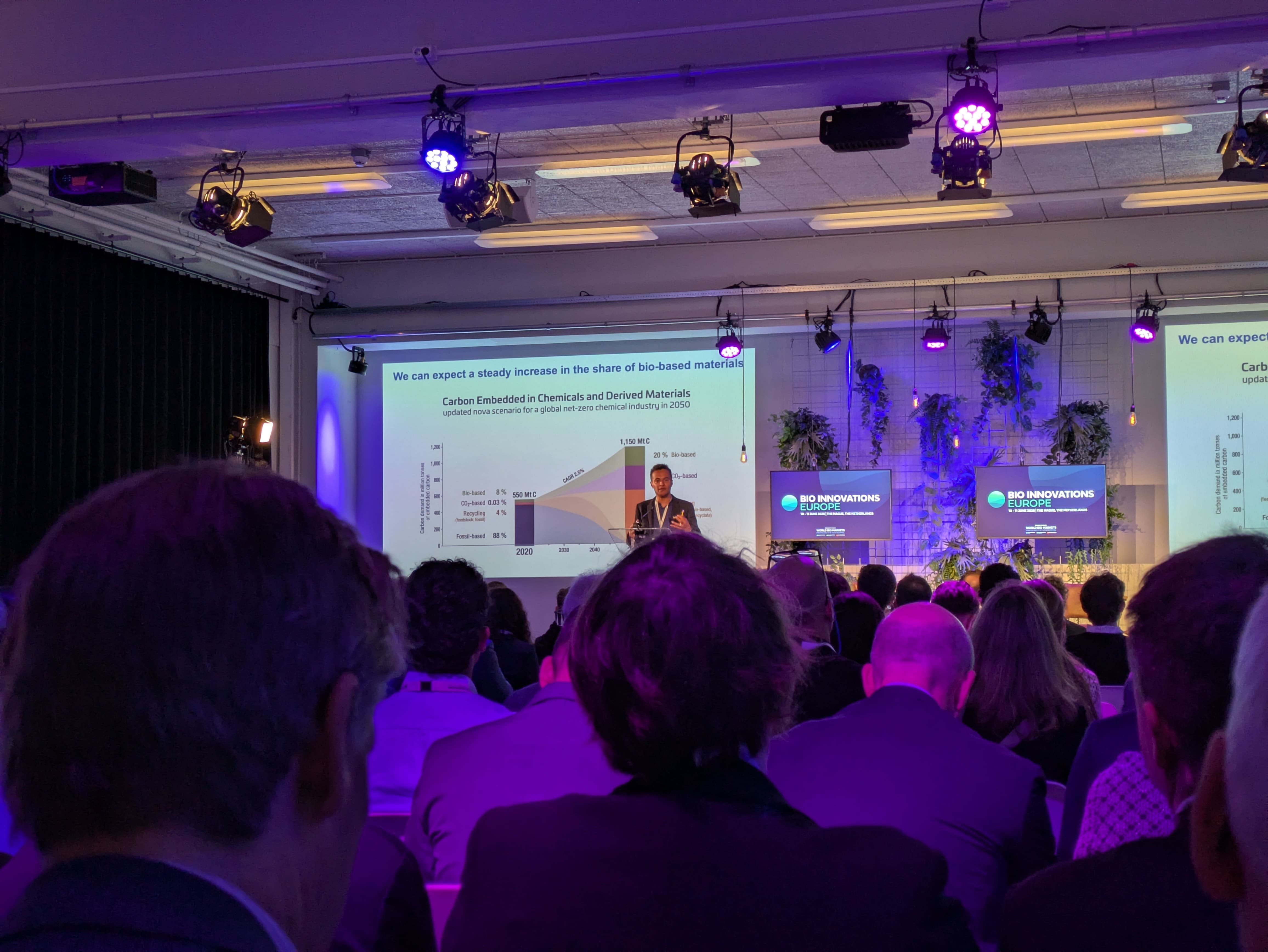 A speaker stands on a lit stage in front of a presentation slide featuring diagrams on the topic of bio-based materials. Next to the stage are monitors displaying the “Bio Innovations Europe” logo. In the foreground, the audience sits in a dimly lit room, looking toward the stage.