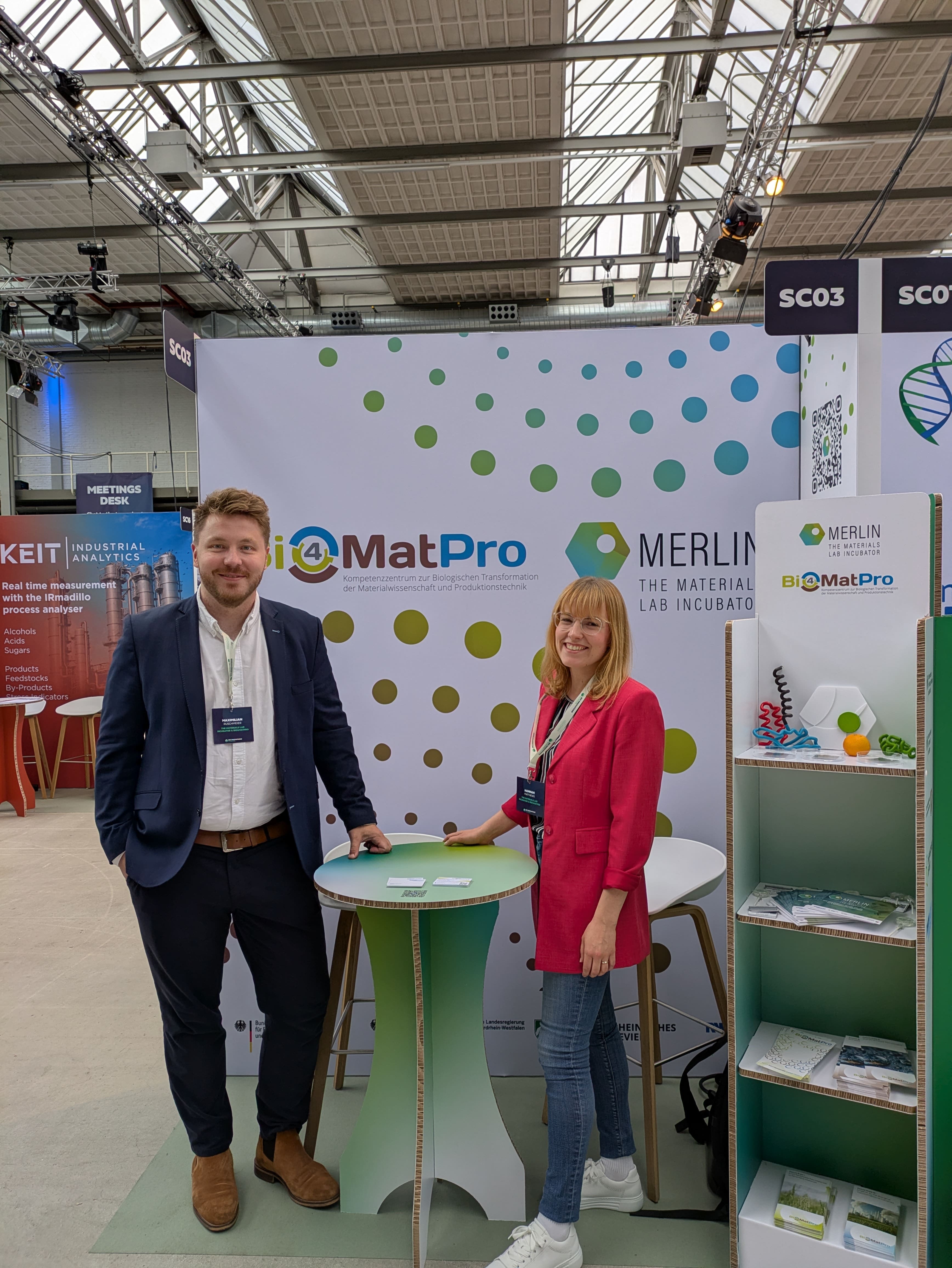 A man in a dark suit and a woman in a bright pink jacket are smiling at a bar table on a trade show booth. The booth prominently displays the BioMatPro and MERLIN The Materials Lab Incubator logos. To the right is a cardboard shelf with informational materials and product displays.