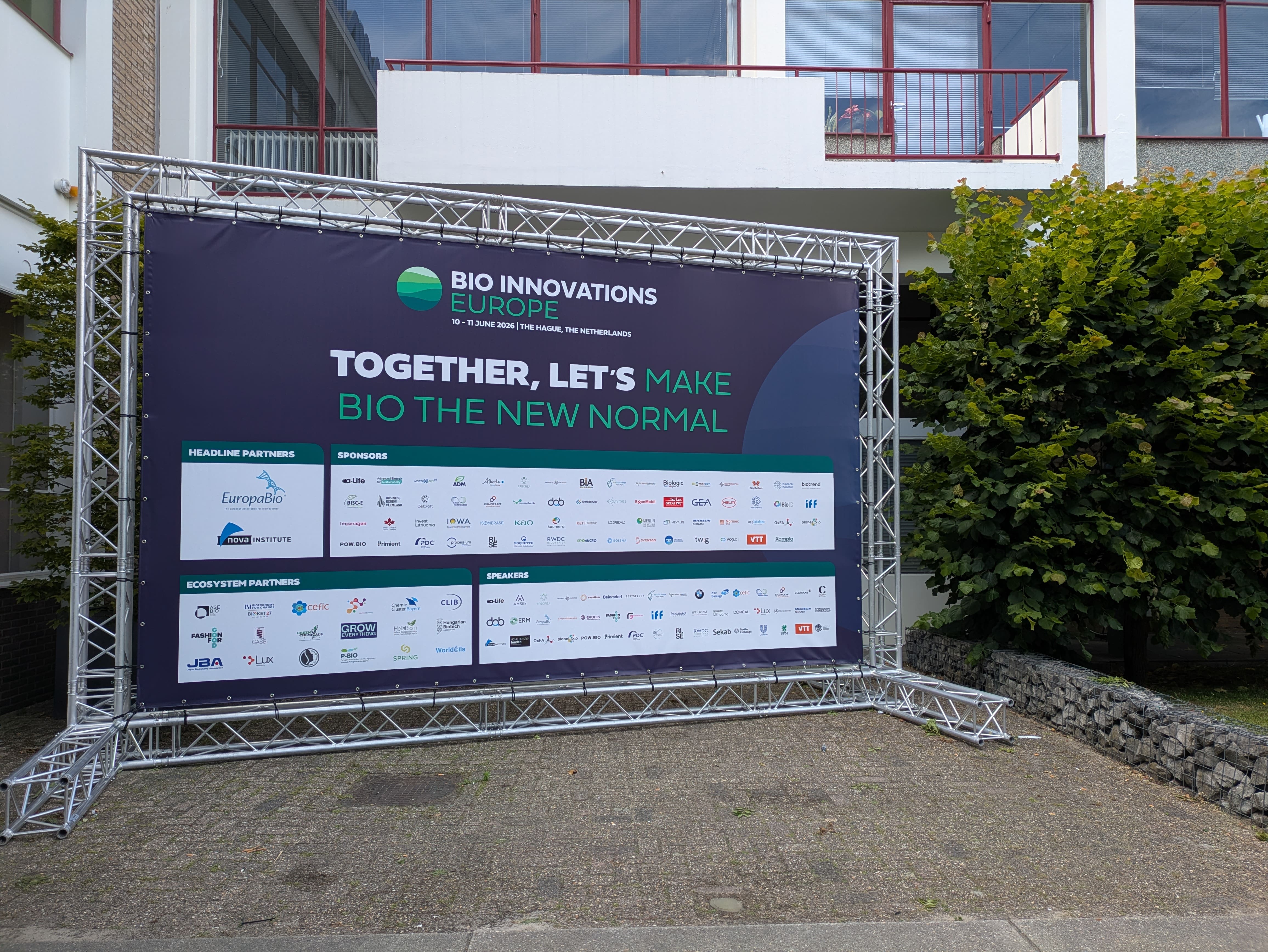 A large, dark event banner bearing the slogan “Bio Innovations Europe – Together, let’s make bio the new normal” is mounted on a metal truss frame in the outdoor area. The banner features numerous logos of partners, sponsors, and speakers. In the background is a building with red windows and greenery.