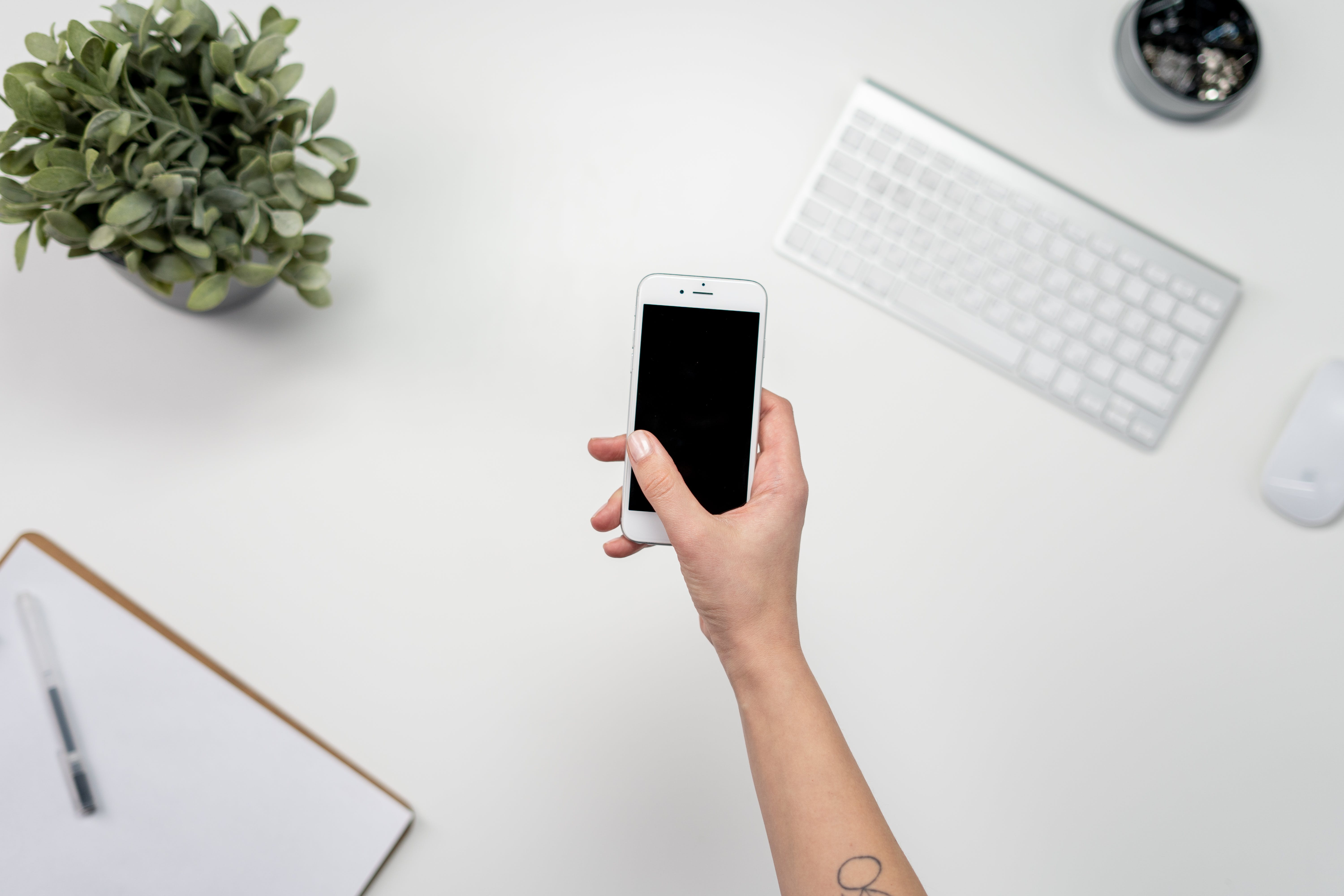 Eine Person hält ein Mobiltelefon in der Hand. Die Hand wird über einen Schreibtisch gehalten.